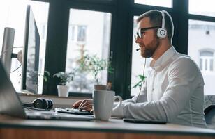 In the call center. Wearing ,headphones. Office worker in formal clothes is sitting by computer and working photo