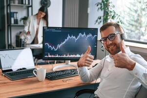 Man showing thumbs up. Graphs on the screen. Two employees are working together in the modern office photo