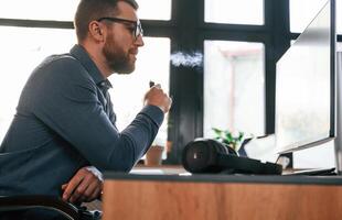 Smoking, electric cigarette. Man in formal clothes is working in the modern office. Using computer photo