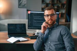 Software, developer is sitting, and posing. Man in formal clothes is working in the modern office. Using computer photo