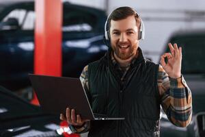 Smiling, standing, holding laptop. In headphones. Man in uniform is working in the auto salon photo