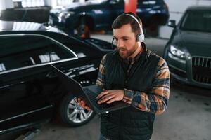 Standing with laptop in hands and in headphones. Man in uniform is working in the auto salon photo