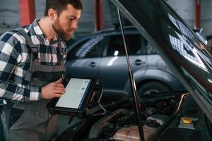 Standing near, the car with hood opened. Holding tablet. Man in uniform is working in the auto salon photo