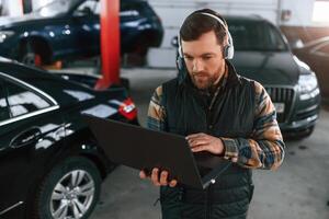Standing ,with, laptop in hands and in headphones. Man in uniform is working in the auto salon photo