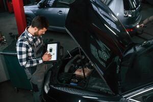 Standing, near the car with hood opened. Holding tablet. Man in uniform is working in the auto salon photo