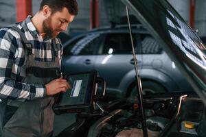 Standing ,near the, car with hood opened. Holding tablet. Man in uniform is working in the auto salon photo