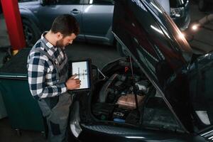 Standing near the car with hood opened. Holding tablet. Man in uniform is working in the auto salon photo