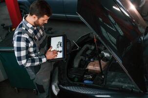 Standing ,,near the car with hood opened. Holding tablet. Man in uniform is working in the auto salon photo