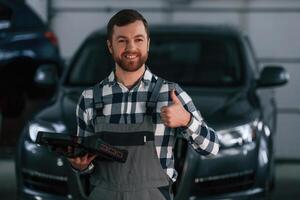 Showing, gesture and smiling. Holding tablet. Man in uniform is working in the auto salon photo