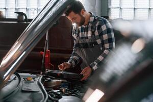 Hood ,,of the car is opened. Using tablet. Man in uniform is working in the auto salon photo
