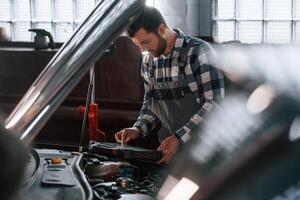 Hood of ,the car is opened. Using tablet. Man in uniform is working in the auto salon photo