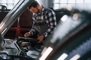 Hood of, the, car is opened. Using tablet. Man in uniform is working in the auto salon photo