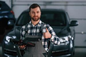 Showing gesture and smiling. Holding tablet. Man in uniform is working in the auto salon photo