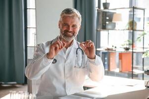 Sitting and smiling. Senior doctor in white coat,is at work indoors photo