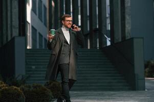 Amazing sunlight, holding, smartphone and cup of drink. Handsome man in formal clothes is outdoor near the business building photo