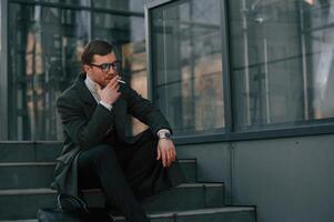Sitting, on the stairs and smoking. Handsome man in formal clothes is outdoor near the business building photo