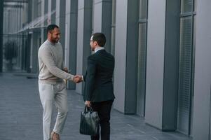 Doing, handshake, greetings gesture. Businessman in formal clothes is with black man outdoors in the city photo