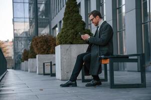 Using ,social media by ,smartphone. Handsome man in formal clothes is outdoor near the business building photo