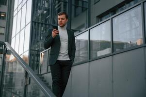 Using ,social media by smartphone. Handsome man in formal clothes is outdoor near the business building photo