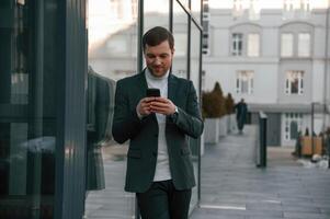 Using social media by smartphone. Handsome man in formal clothes is outdoor near the business building photo