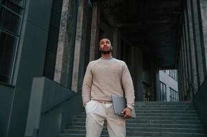 Front view. .Standing ,with laptop in hands. Handsome black man is outdoors near the business building photo