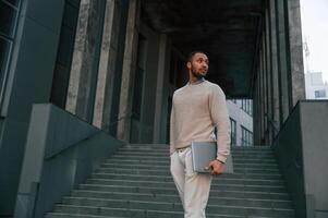 Front view. ,Standing ,with laptop in hands. Handsome black man is outdoors near the business building photo