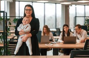 Happy mother is ,standing and holding her little son. Infant baby is in the office where group of people are working together photo