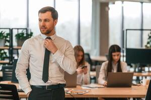 Man, in tie is standing in front of his colleagues. Business people in formal clothes are working in the office together photo