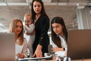 Woman is standing with son in hands.. Infant baby is in the office where group of people are working together photo