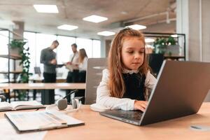 Sitting ,by the ,table with laptop. Little girl is in the office where people working together photo