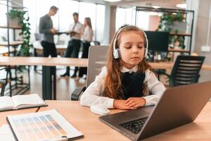 Sitting by the table with laptop. Little girl is in ,the, office where people working together photo