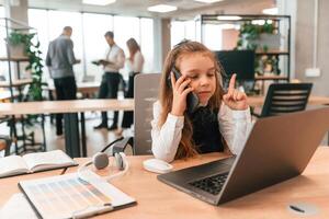 Sitting, by the table, with laptop. Little girl is in the office where people working together photo