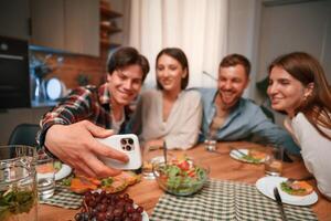 Man is making selfie. Group of people are on the kitchen, having dinner, spending time together photo