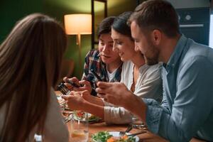 Sitting by the table, with smartphones in hands. Group of people are on the kitchen, having dinner, spending time together photo