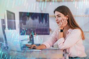 Graphs, data, different finance information. Using laptop. Female stock broker in casual clothes is working in the office by pc photo