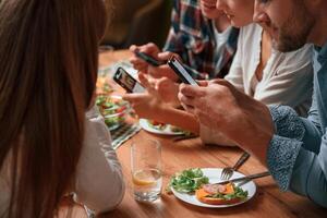 Sitting by, the table, with smartphones in hands. Group of people are on the kitchen, having dinner, spending time together photo
