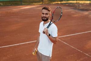 Positive young man is on the tennis court at ,sunny daytime. holding racket photo