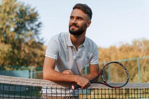 Standing with ,racket in hands. Young man is on the tennis court at sunny daytime photo