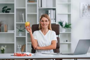 Smiling, holding an apple. Young female doctor in white coat is indoors photo