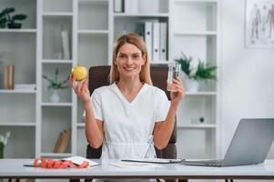 Healthy, eating, holding an apple and glass of water. Conception of diet. Young female doctor in white coat is indoors photo