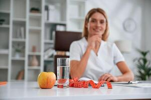 An apple and measuring ,tape are on the table.. Young female doctor in white coat is indoors photo