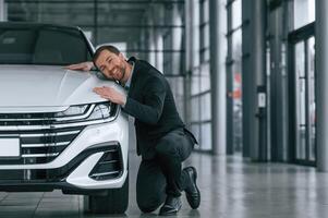 Embracing the ,vehicle. Man in black suit is indoors in the car dealership near automobile photo