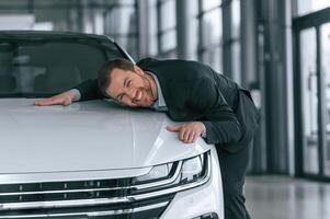 Embracing, the vehicle. Man in black suit is indoors in the car dealership near automobile photo