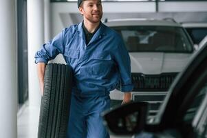 Walking, forward ,and holding the tire. Man in blue uniform is working in the car service photo