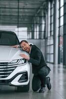 Embracing the vehicle. Man in black suit is indoors in the car dealership near automobile photo