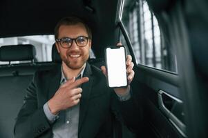 Holding smartphone. Man in formal business clothes is sitting in the modern automobile photo