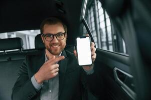 Holding ,smartphone. Man in formal business clothes is sitting in the modern automobile photo
