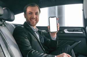 Holding smartphone. Man in formal business ,,clothes is sitting in the modern automobile photo