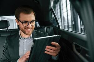 Smiling, using, tablet. Man in formal business clothes is sitting in the modern automobile photo