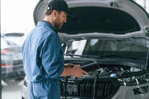 Holding, electricity measuring tool. Man in blue uniform is working in the car service photo